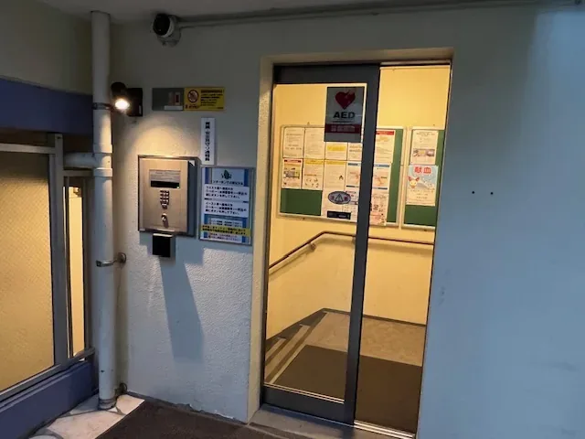 イースト棟の玄関。脇にインターホンと呼出方法の案内、扉の向こうにAEDの表示と掲示板があります。