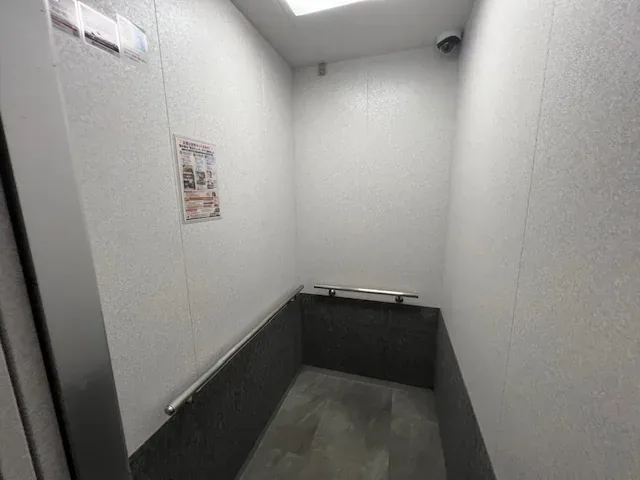 エレベーターの中。白い壁に手すりが付き、天井の照明で明るくなっています。