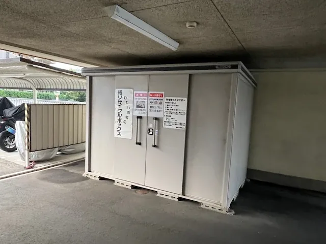 屋根の下に置かれたリサイクルボックス。白い箱型で、扉に案内が貼られています。